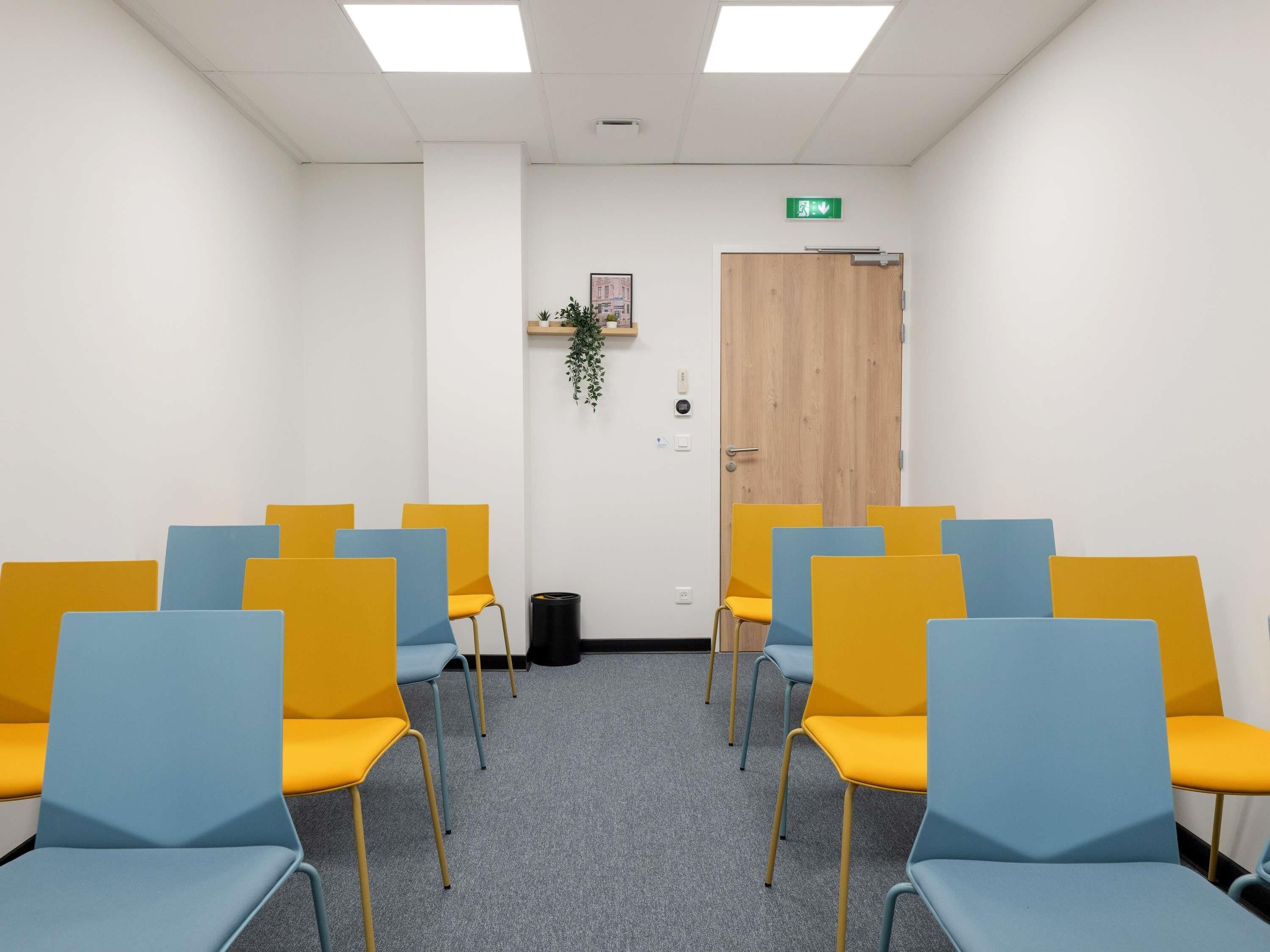 Une pièce avec des rangées de chaises, une porte, une plante et un interrupteur sur le mur.