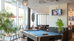 A pool table in a well-lit indoor space with large windows, seating areas, and a television mounted on the wall. - ibis Lyon Caluire Cité Internationale (Caluire-et-Cuire)