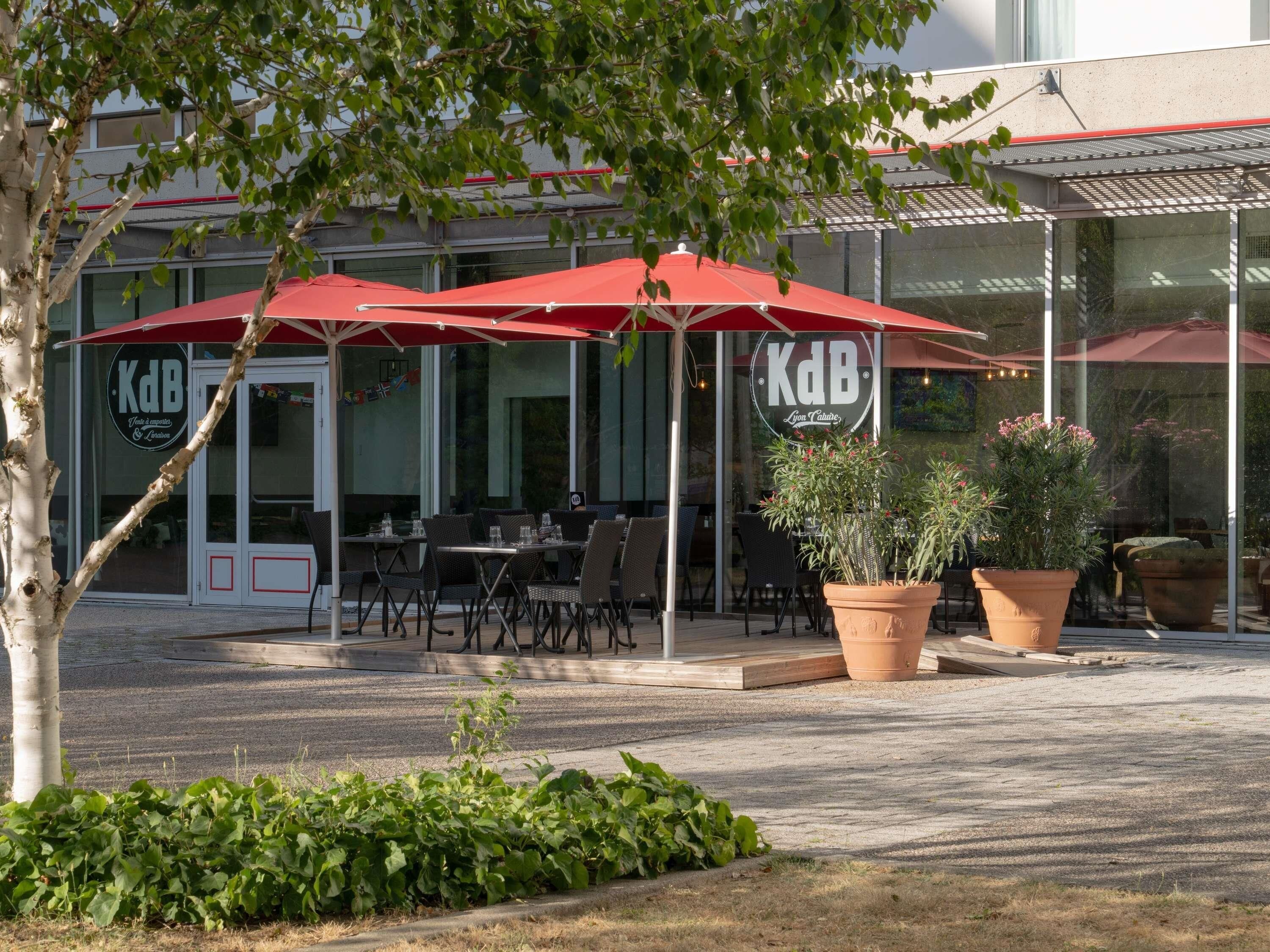 An outdoor seating area with red umbrellas, a restaurant named 'KdB,' and potted plants.