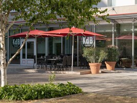 Un área de estar al aire libre con sombrillas rojas, un restaurante llamado 'KdB' y plantas en macetas.