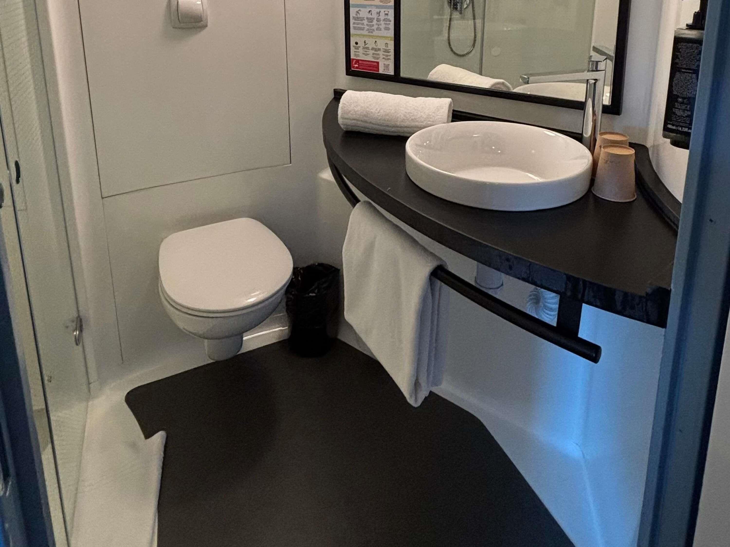A bathroom with a toilet, a sink, a mirror, and a black trash bin.