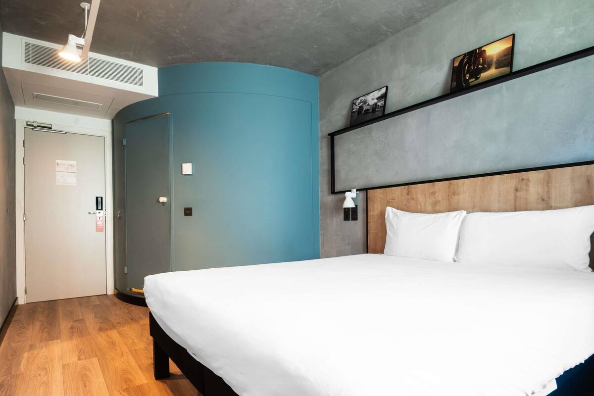A hotel room with a bed, a wooden headboard, a blue accent wall, a door, and a wall-mounted shelf with pictures.