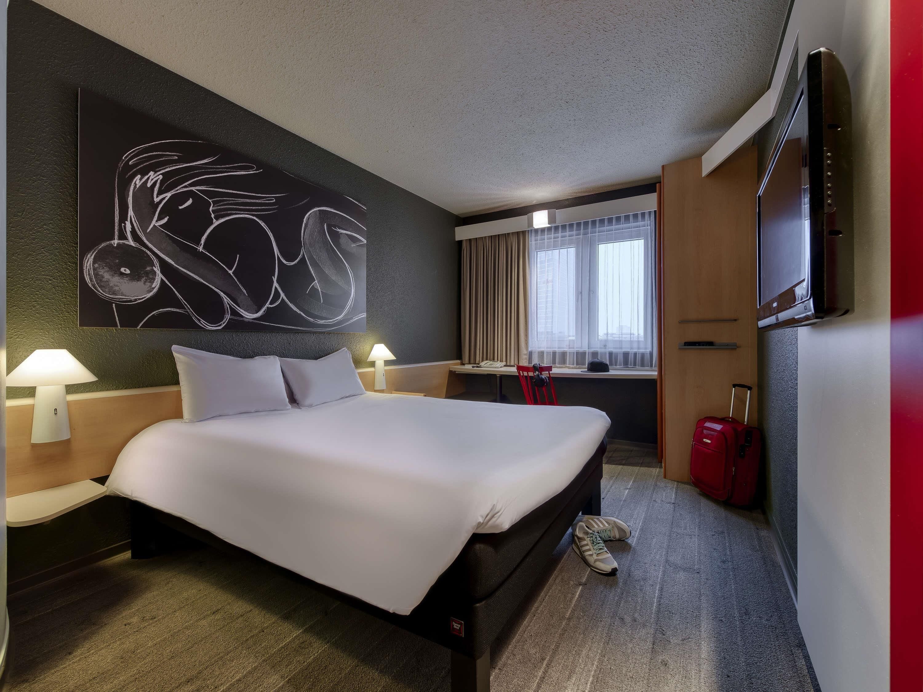 A hotel room with a large bed, two bedside lamps, a desk with a chair, a TV, and a window with curtains.