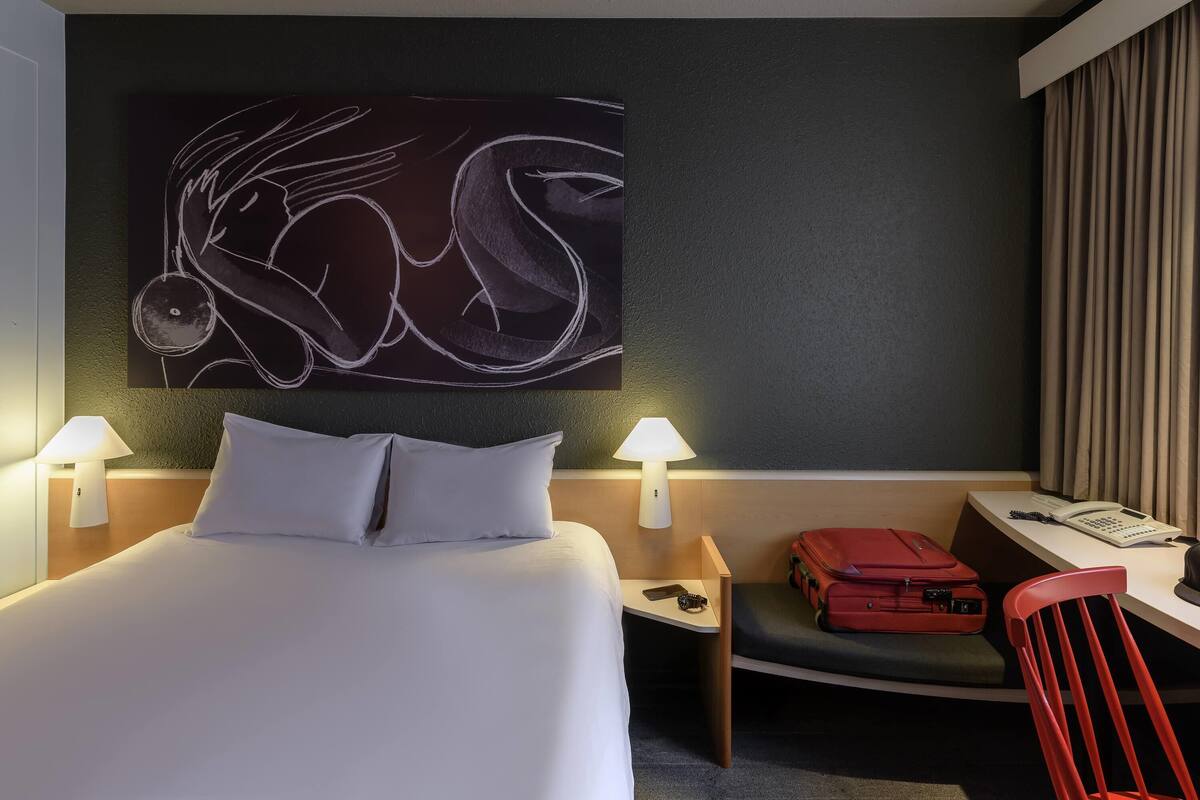 Een hotelkamer met een groot bed, twee bedlampen, een bureau met telefoon, een rode koffer en een stoel.