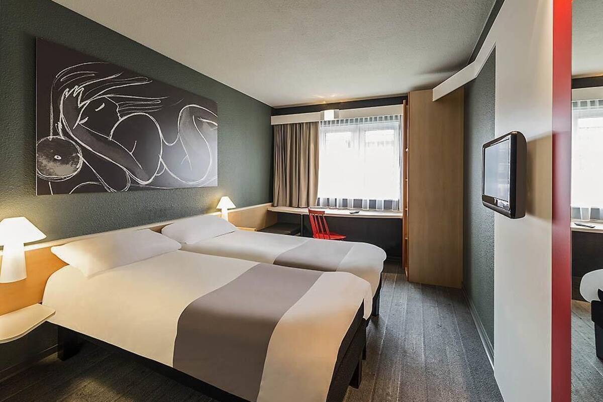 Een hotelkamer met twee bedden, een bureau, een stoel, een televisie en een groot abstract schilderij aan de muur.