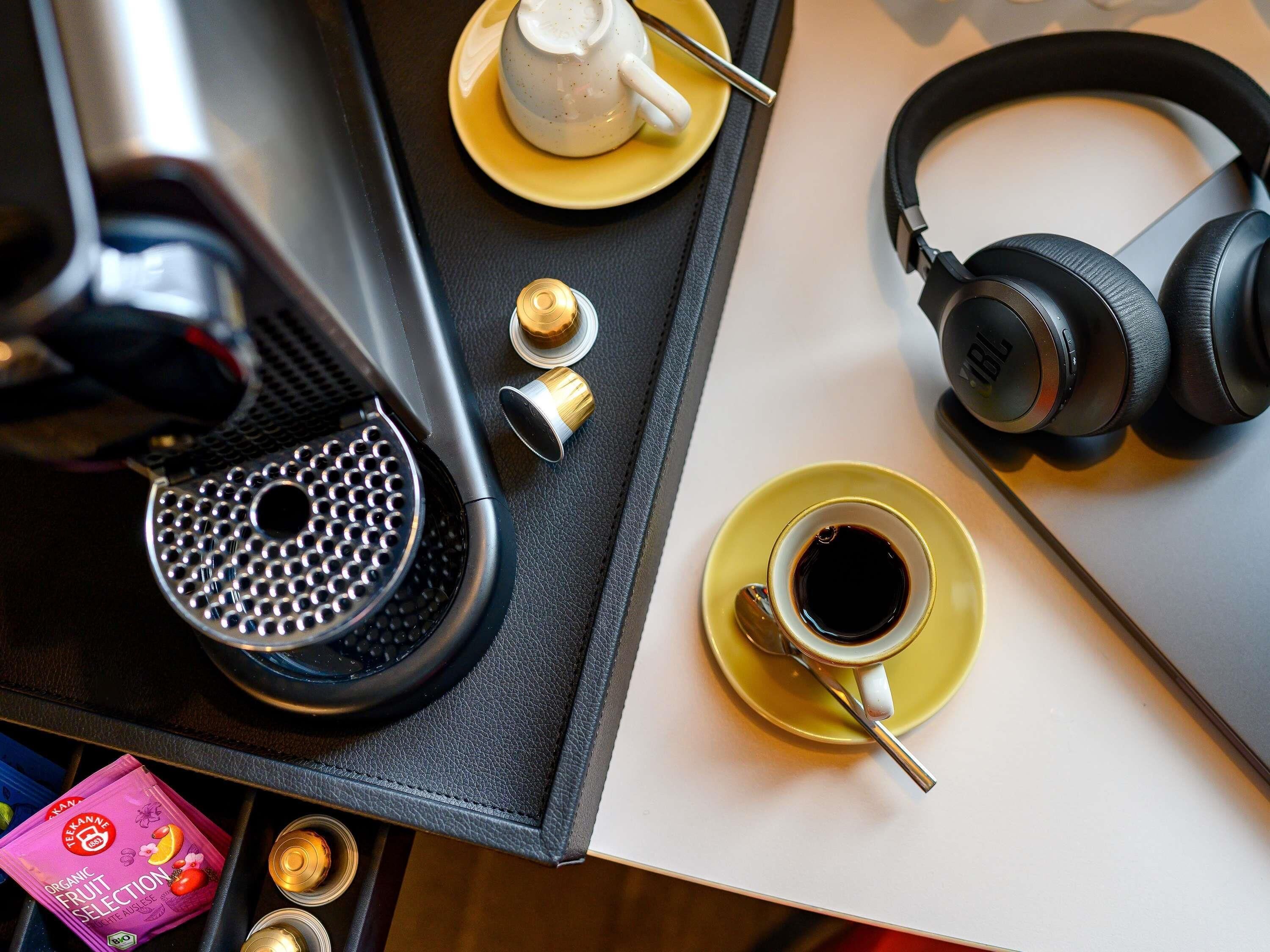 A coffee machine, a cup of coffee, a pair of headphones, and a box of organic fruit selection on a table.