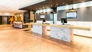 Un espace d’accueil moderne, doté d’un long comptoir de réception, de plusieurs luminaires suspendus et d’un écran numérique affichant des informations.