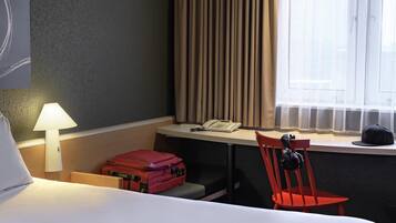 Habitación de hotel con cama, escritorio, teléfono, silla, maleta roja y lámpara.