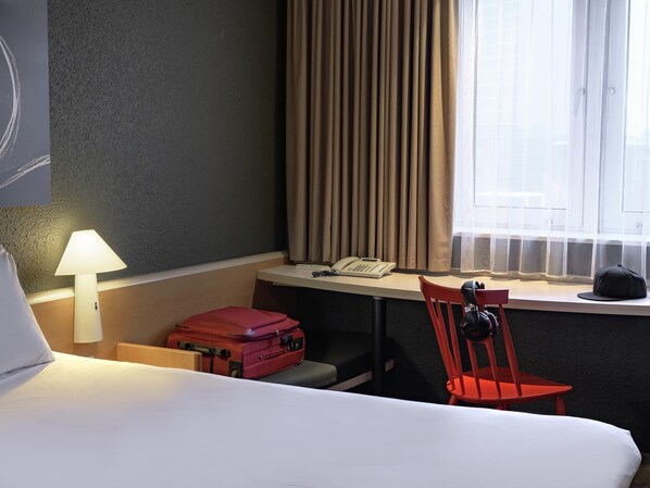 A hotel room with a bed, a desk with a telephone, a chair, a red suitcase, and a lamp.