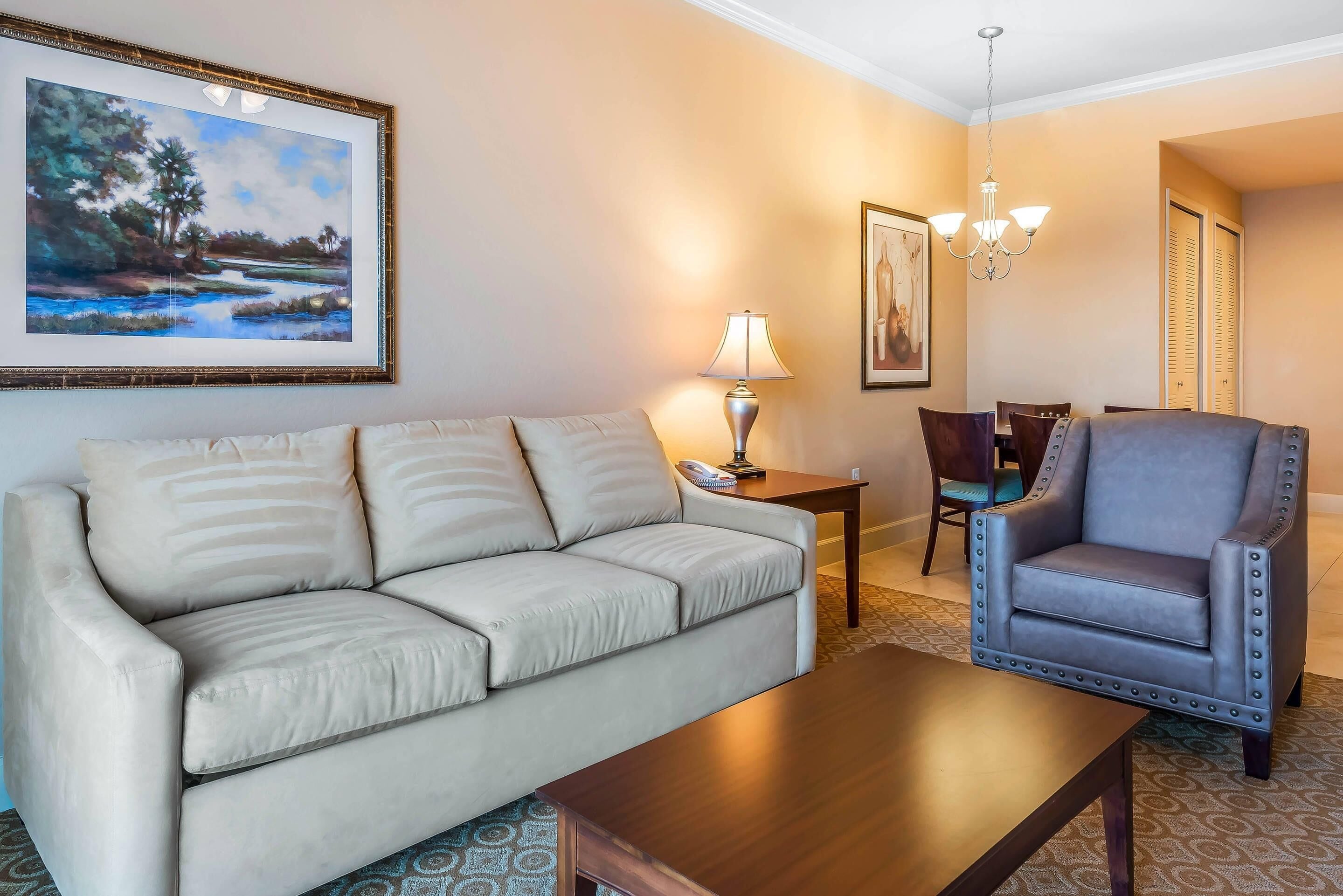 Una sala de estar con un sofá color beige, una mesa de centro de madera, un sillón azul, una lámpara y un cuadro enmarcado en la pared.