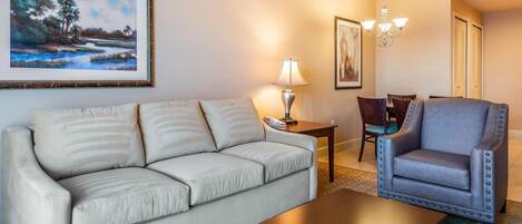 Una sala de estar con un sofá color beige, una mesa de centro de madera, un sillón azul, una lámpara y un cuadro enmarcado en la pared.