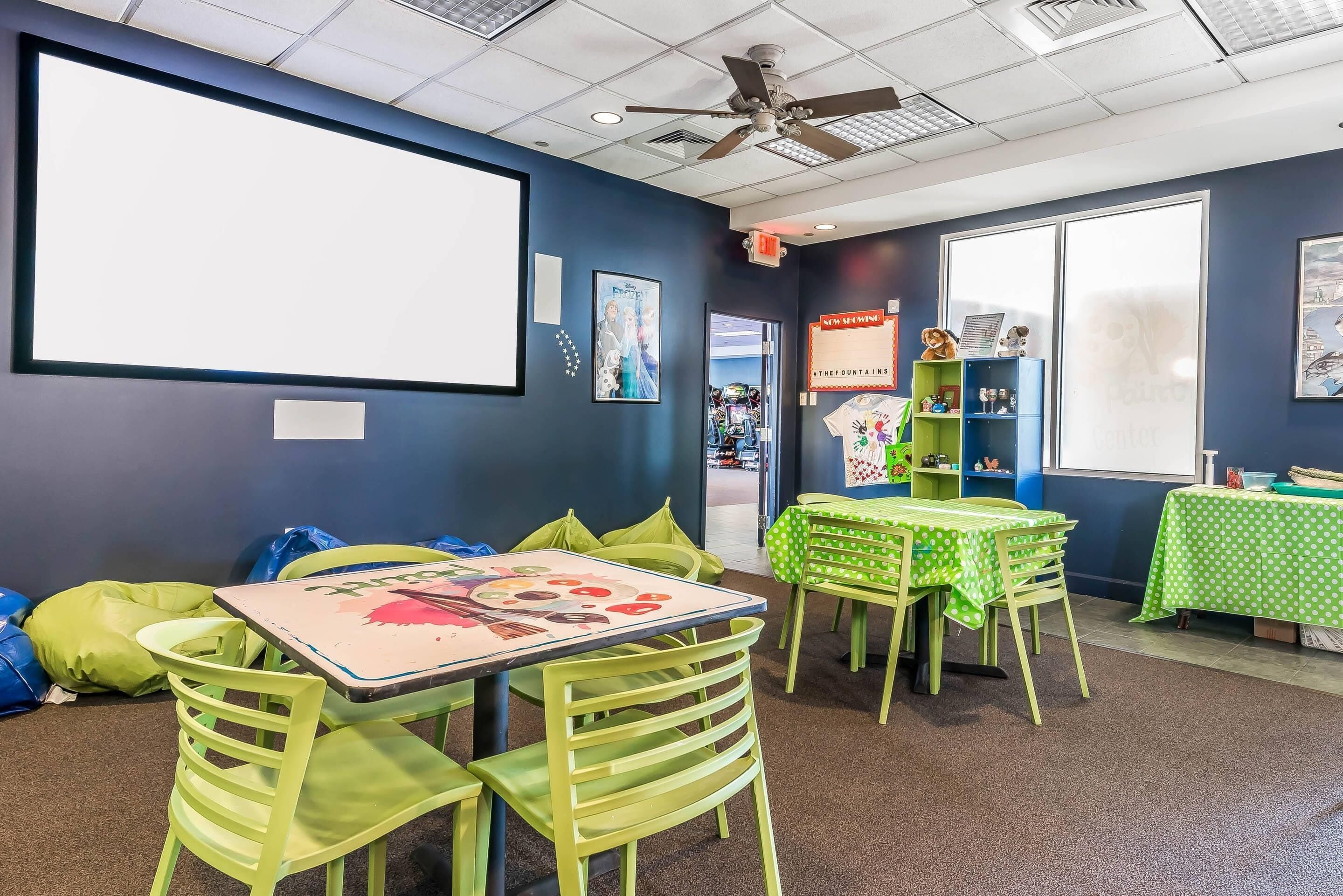 A children's play area with green tables and chairs, a large white screen, and a ceiling fan.
