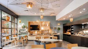 A modern cafe with round tables and chairs, a kitchen area, and a large window with greenery visible outside. - Novotel Suites Cannes Centre (Cannes)