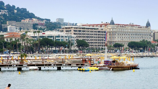 A beach with people sunbathing, a pier with boats, and a large building in the background. - Novotel Suites Cannes Centre (Cannes)