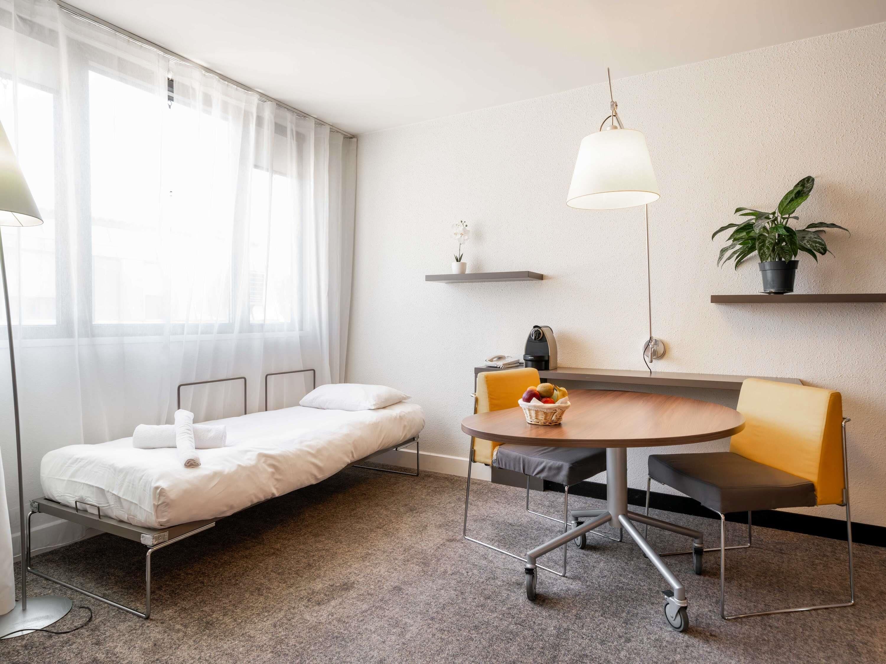 A compact hotel room with a single bed, a small round table with chairs, a desk, a chair, a potted plant, and a hanging light fixture.