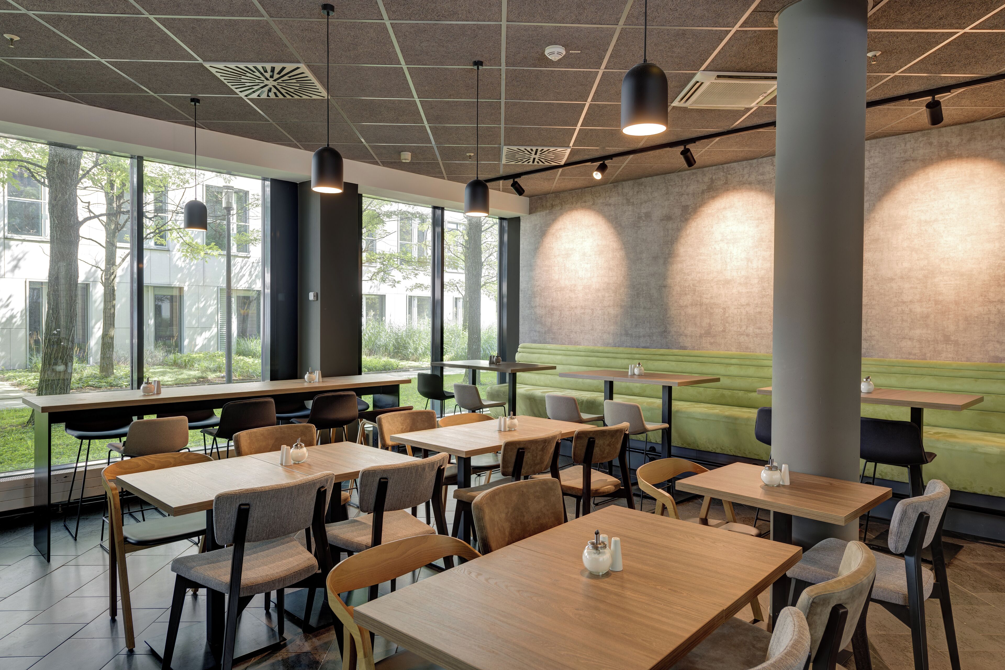 A modern dining area with tables and chairs, large windows, and a view of greenery outside.