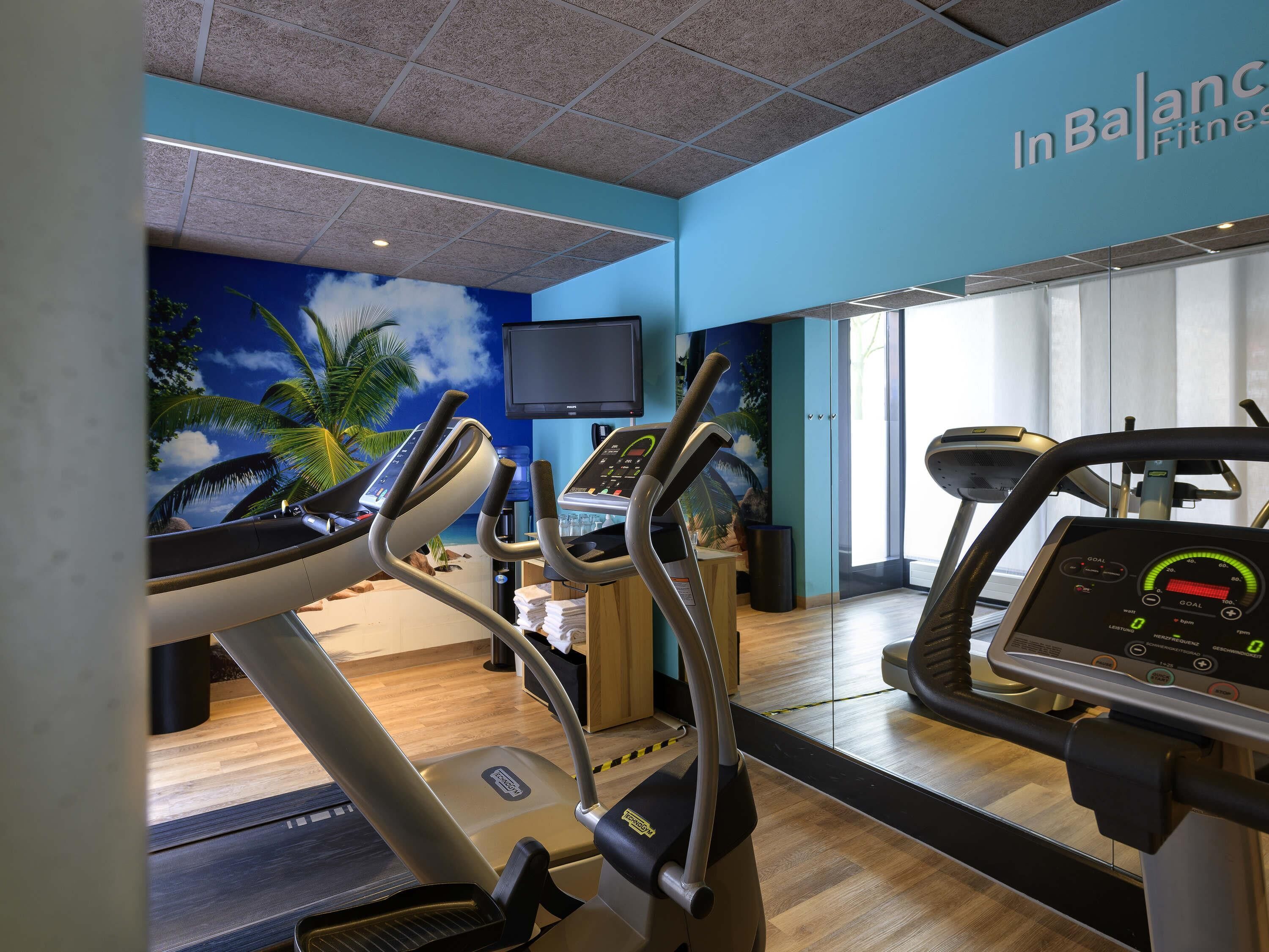 Un gimnasio con cintas de correr, un televisor en la pared y un mural tropical.