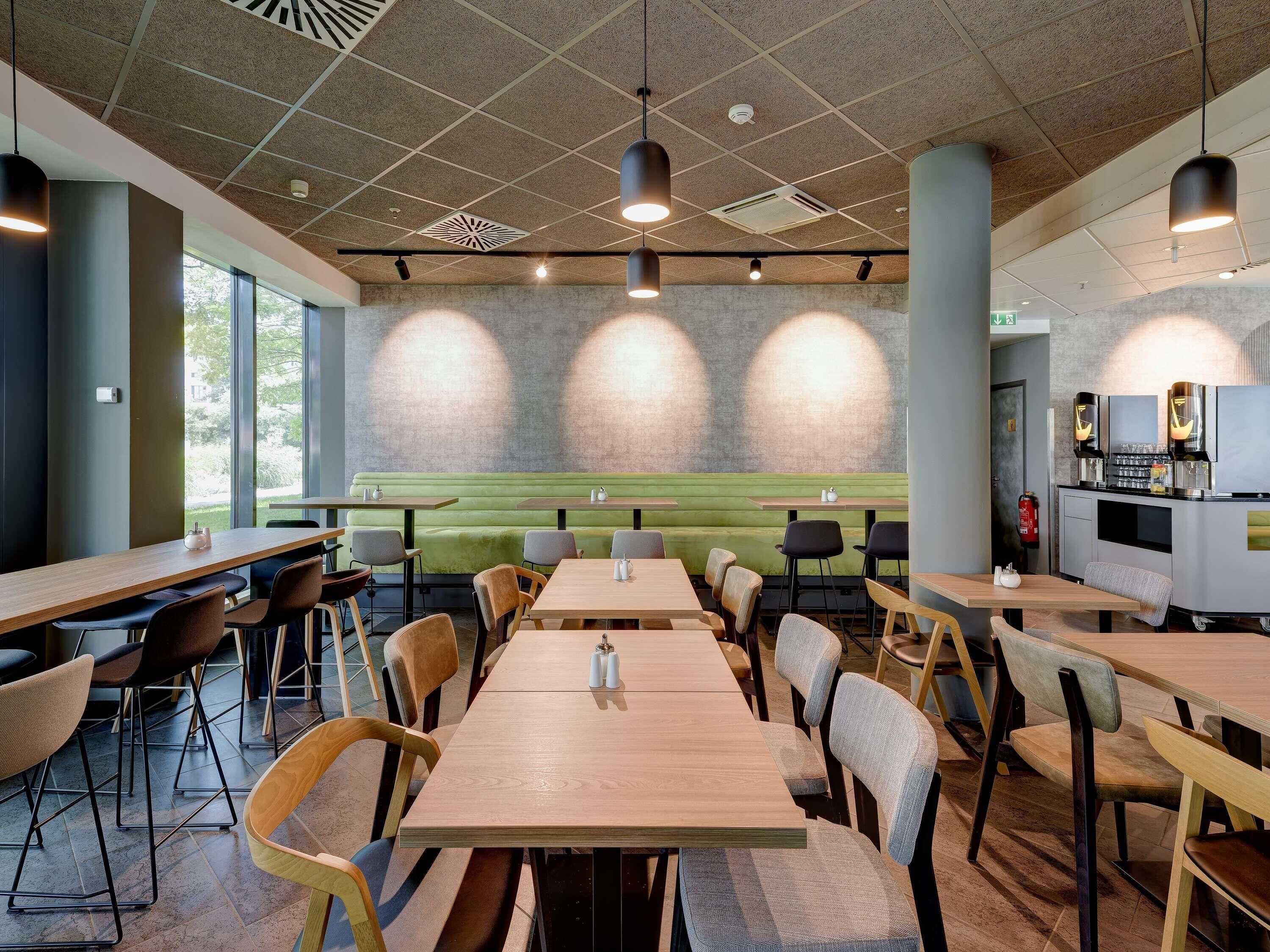 A modern dining area with tables and chairs, a green cushioned bench, and a kitchen counter with a coffee machine.