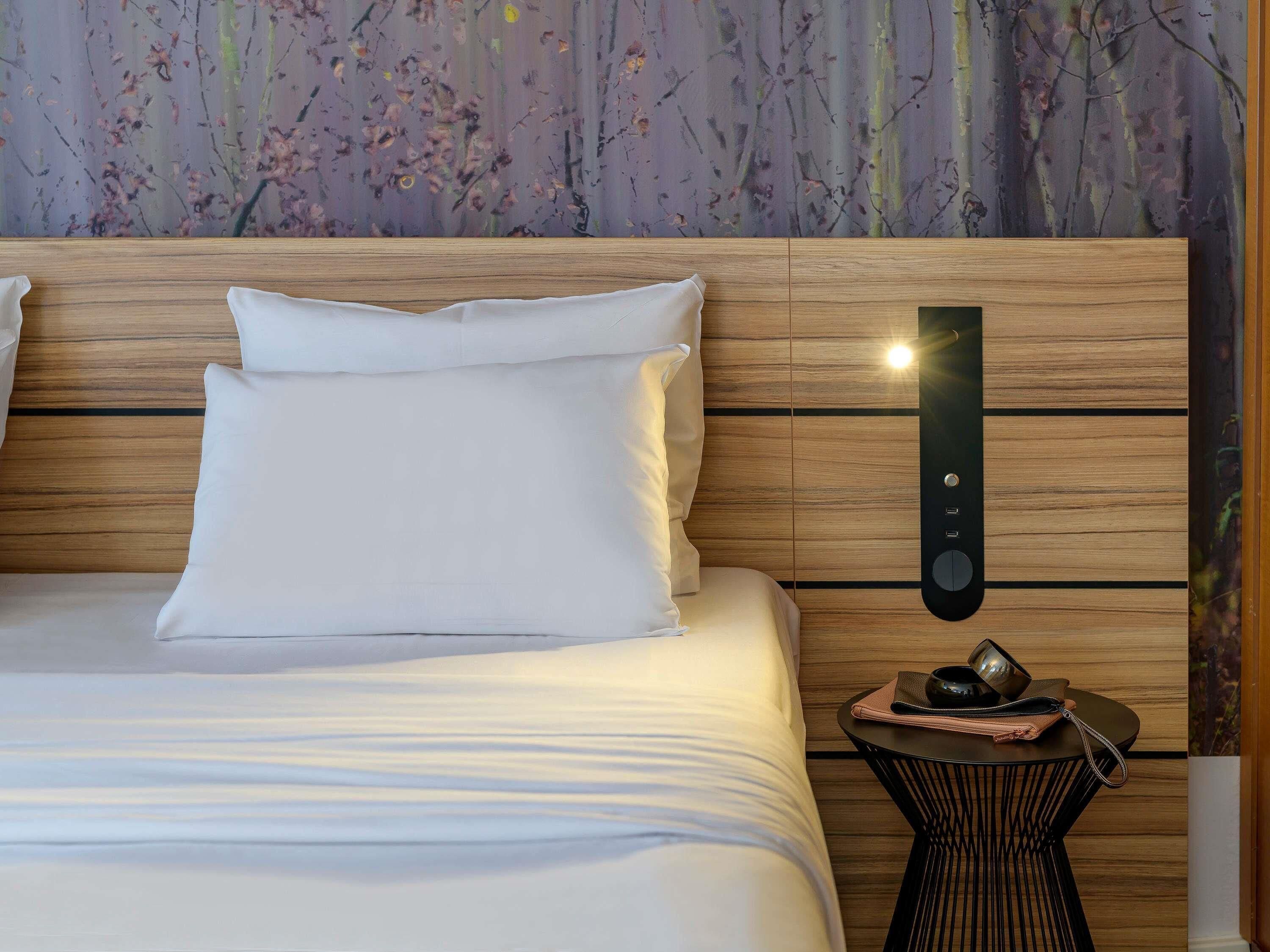 A neatly made bed with white linens and a wooden headboard. A small round table with a black base and a woven top is beside the bed, holding a pair of sunglasses and a small bag.