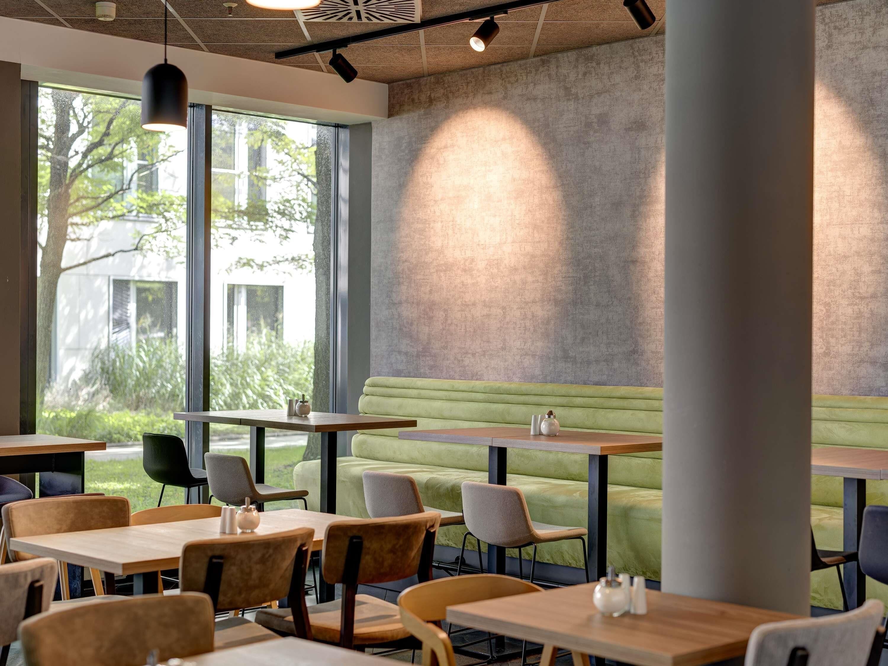 A modern cafe with tables, chairs, a green cushioned bench, and large windows.