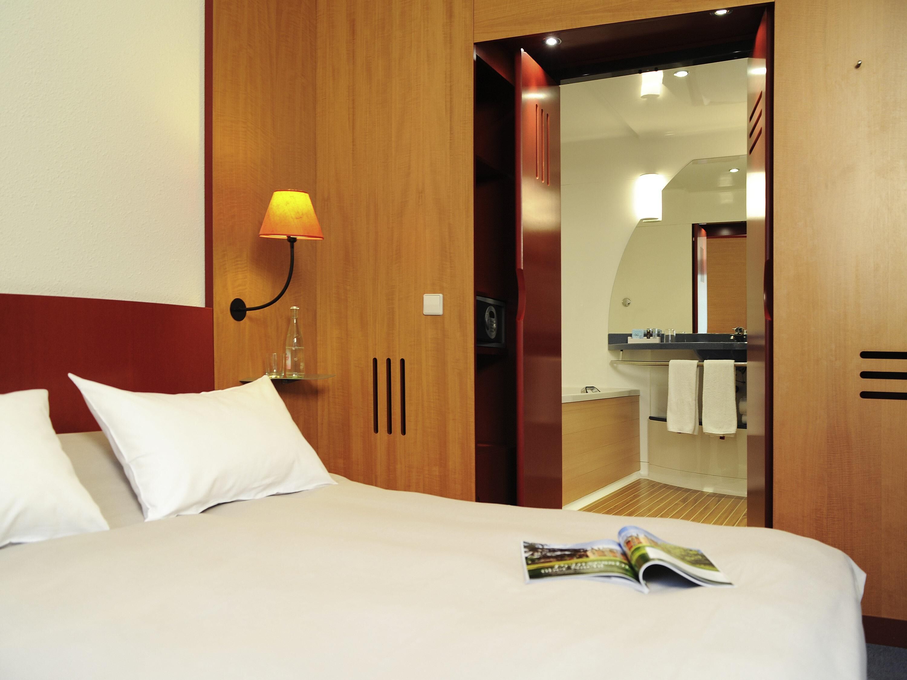 A hotel room with a bed, a nightstand, a lamp, a mirror, and a bathroom visible through an open door.