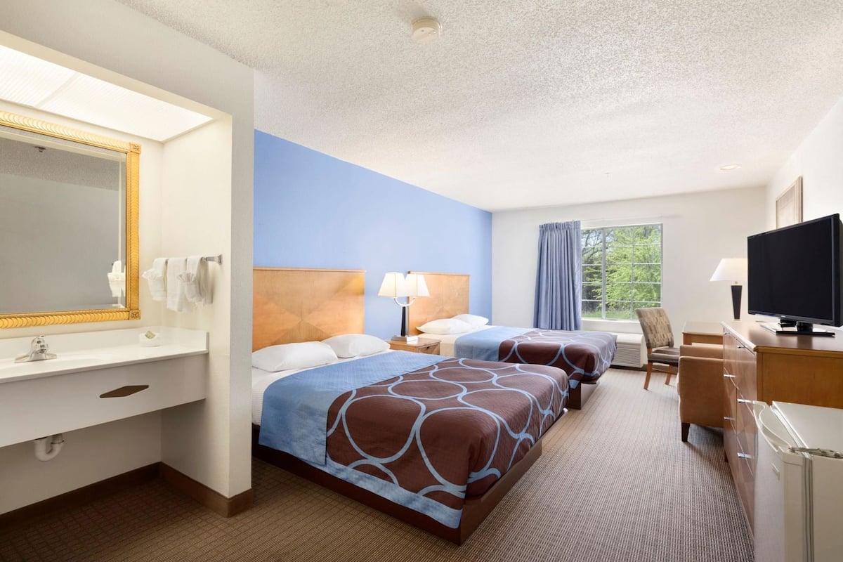 A hotel room with two beds, a desk, a television, and a window with a view of trees.