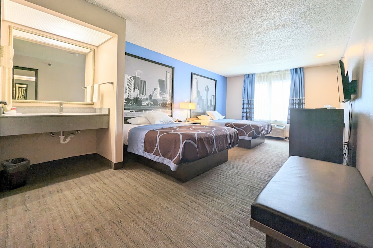 A hotel room with a large bed, a smaller bed, a desk, a mirror, and a cityscape painting on the wall.