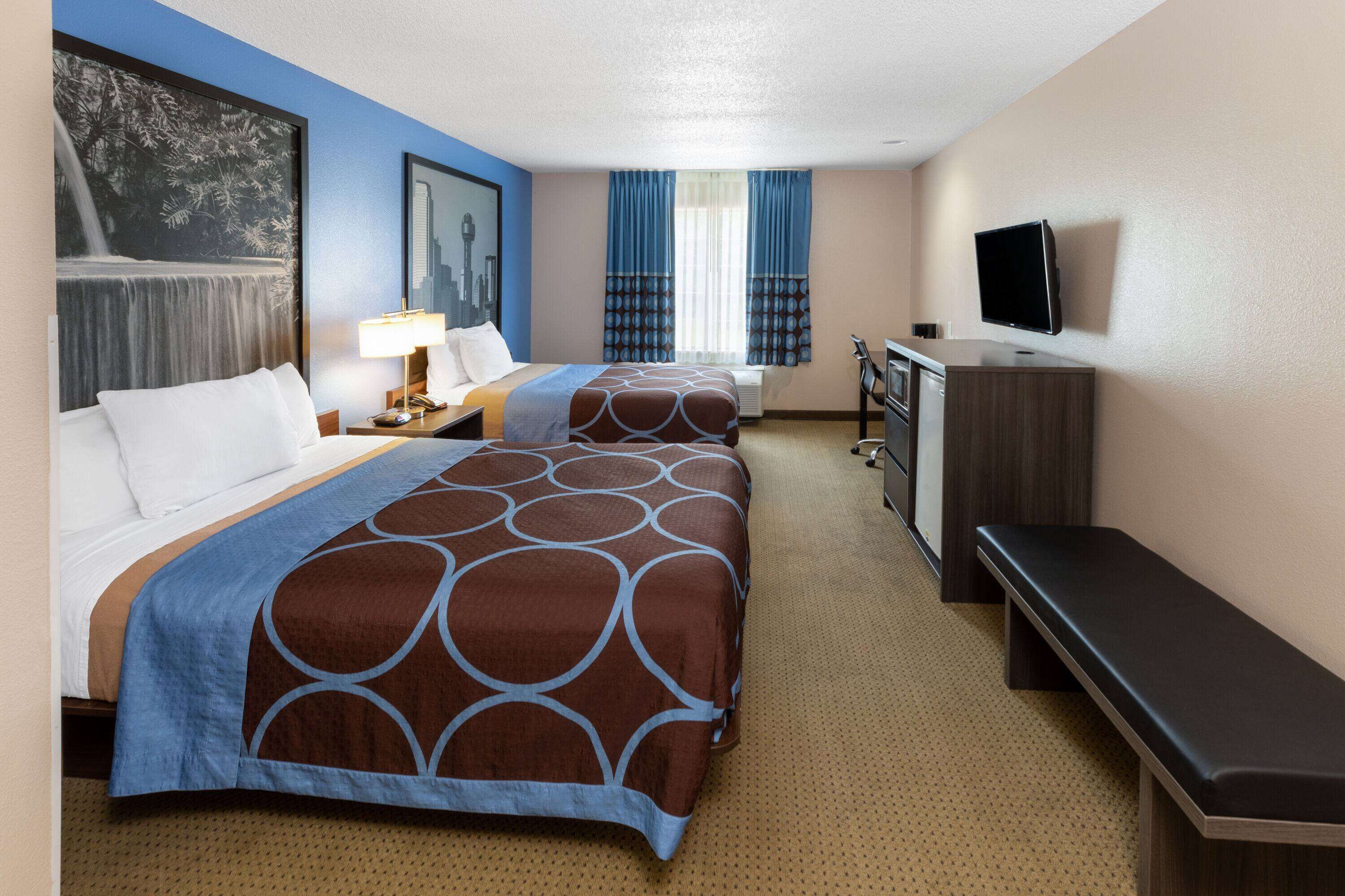 A hotel room with two beds, a desk, a TV, and a large window with curtains.