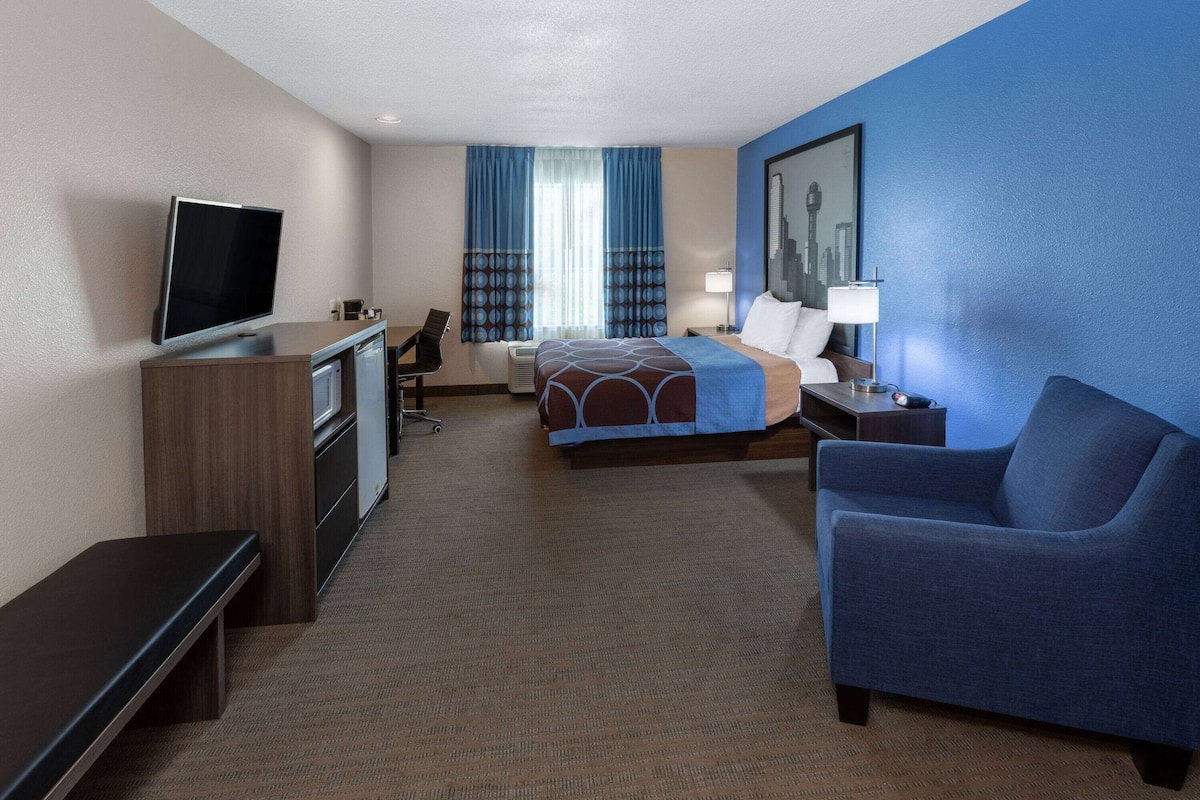 A hotel room with a bed, a desk with a TV, a blue armchair, and a cityscape painting on the wall.