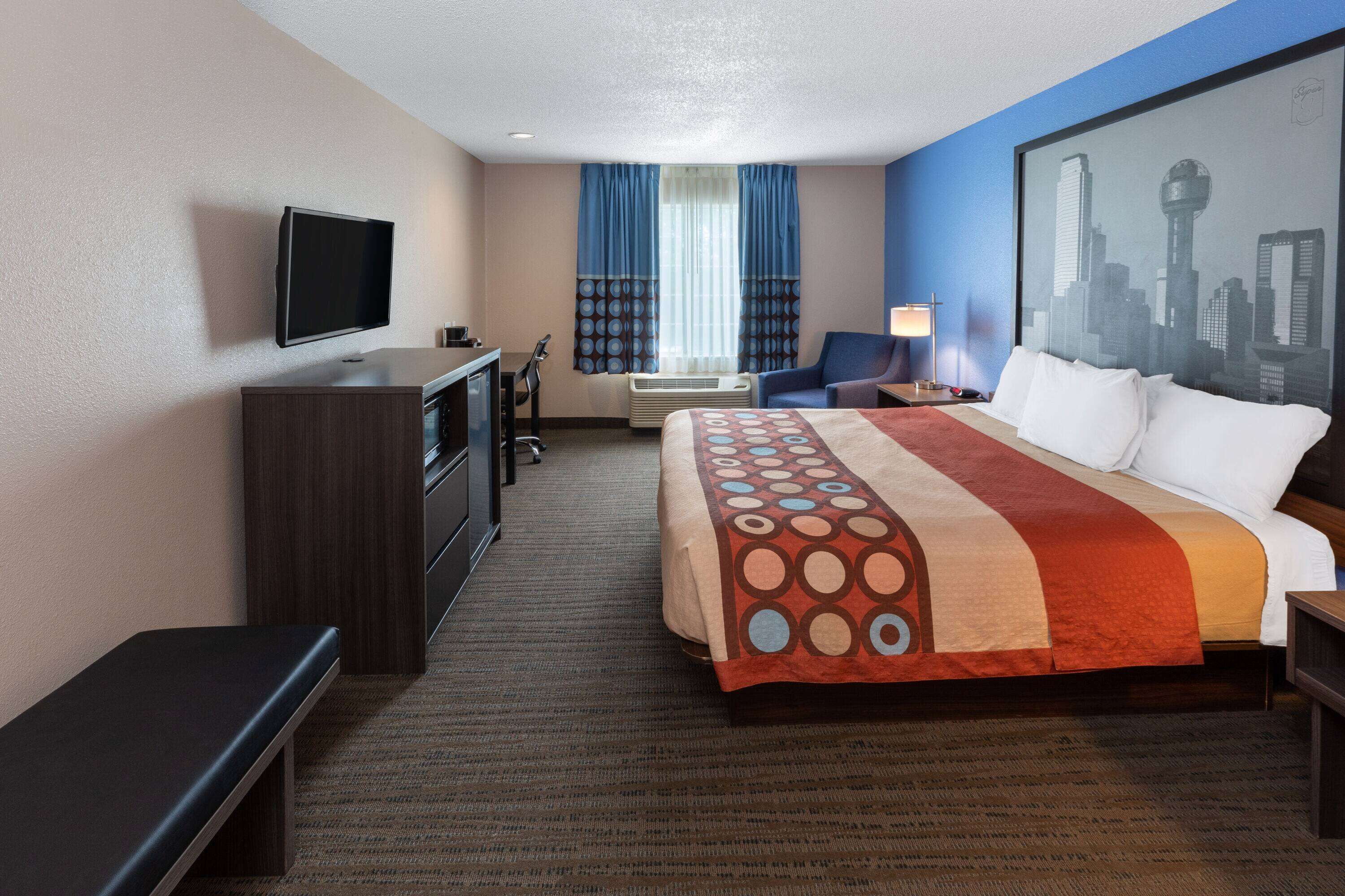 A hotel room with a bed, a desk, a chair, a television, and a cityscape painting on the wall.