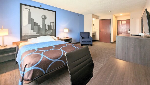 Une chambre d’hôtel avec un lit, une chaise, un bureau, un tableau représentant un paysage urbain et une porte donnant sur une autre pièce.