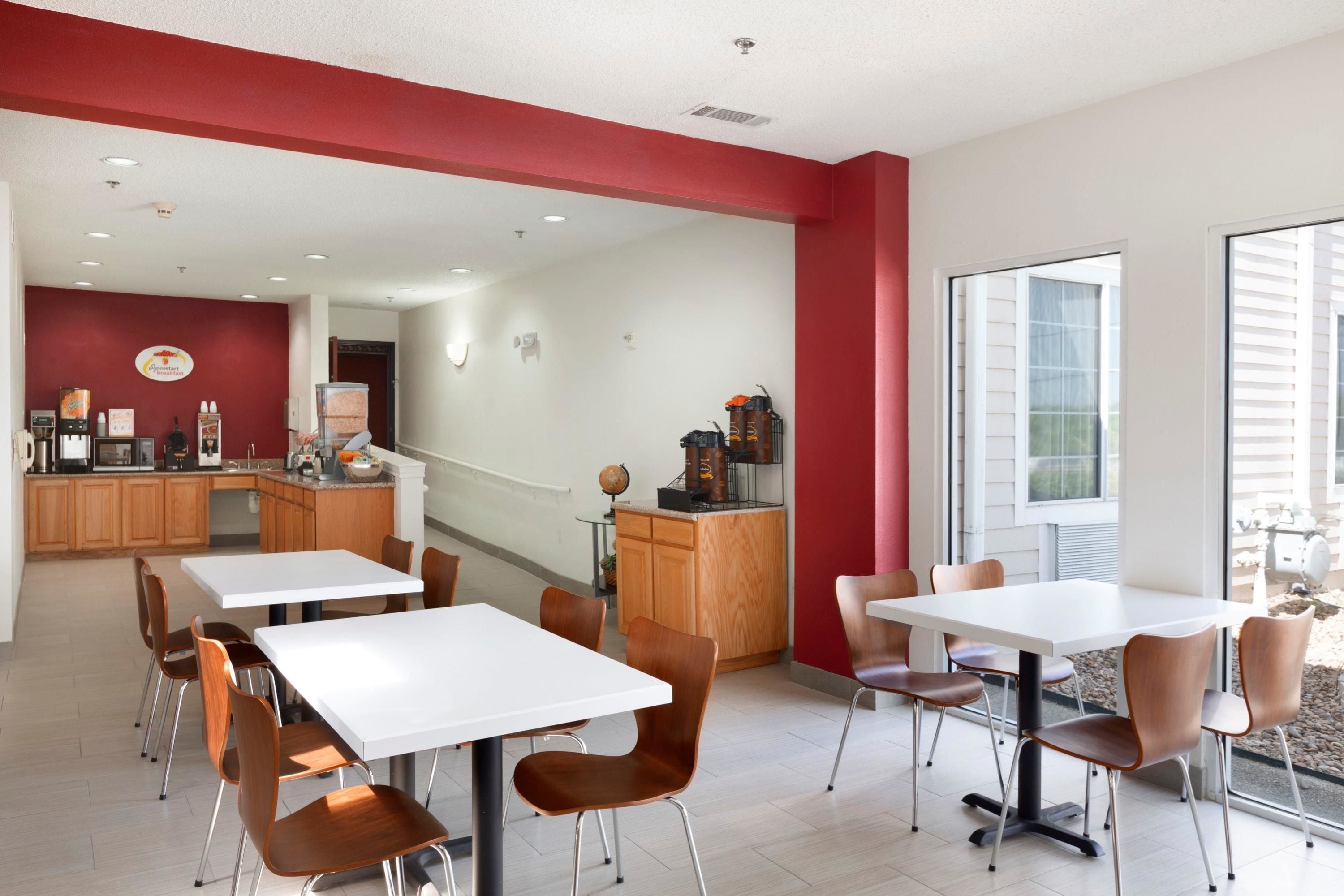 A modern dining area with white tables and brown chairs, a kitchen in the background, and large windows.