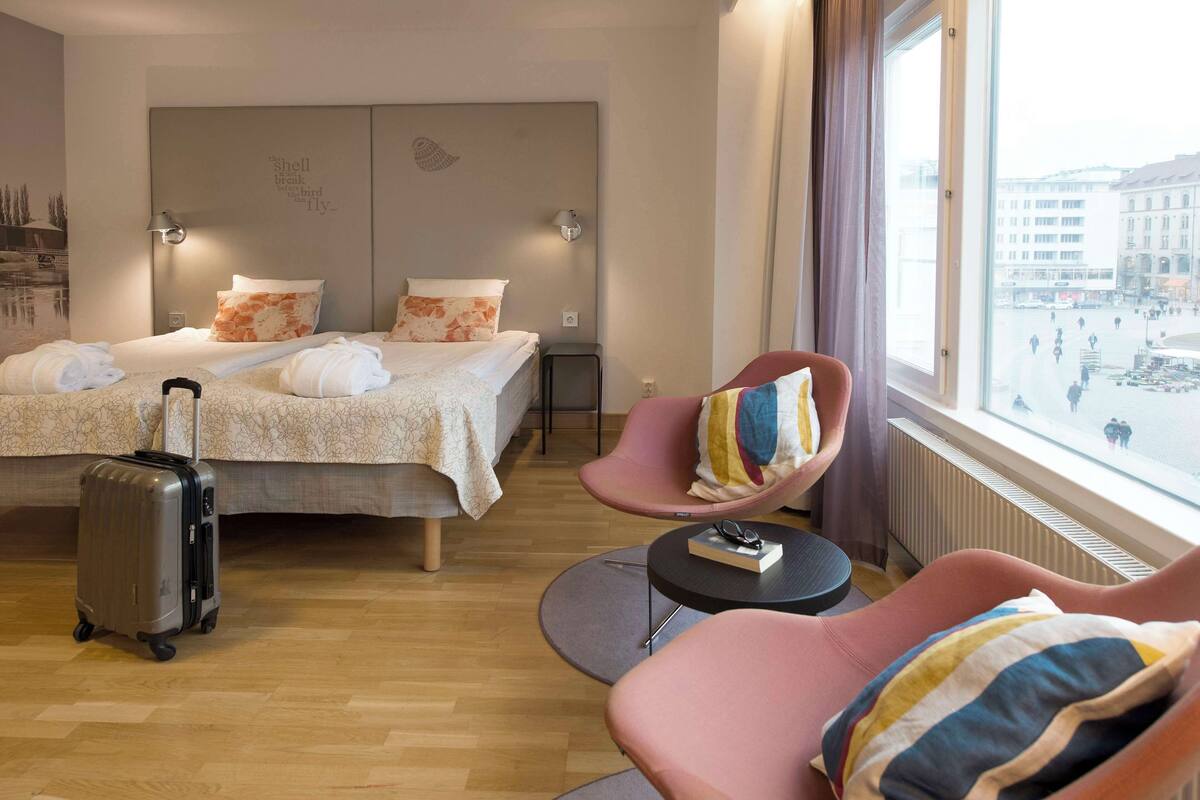 A hotel room with two beds, a sitting area with a chair, a small table, and a suitcase.