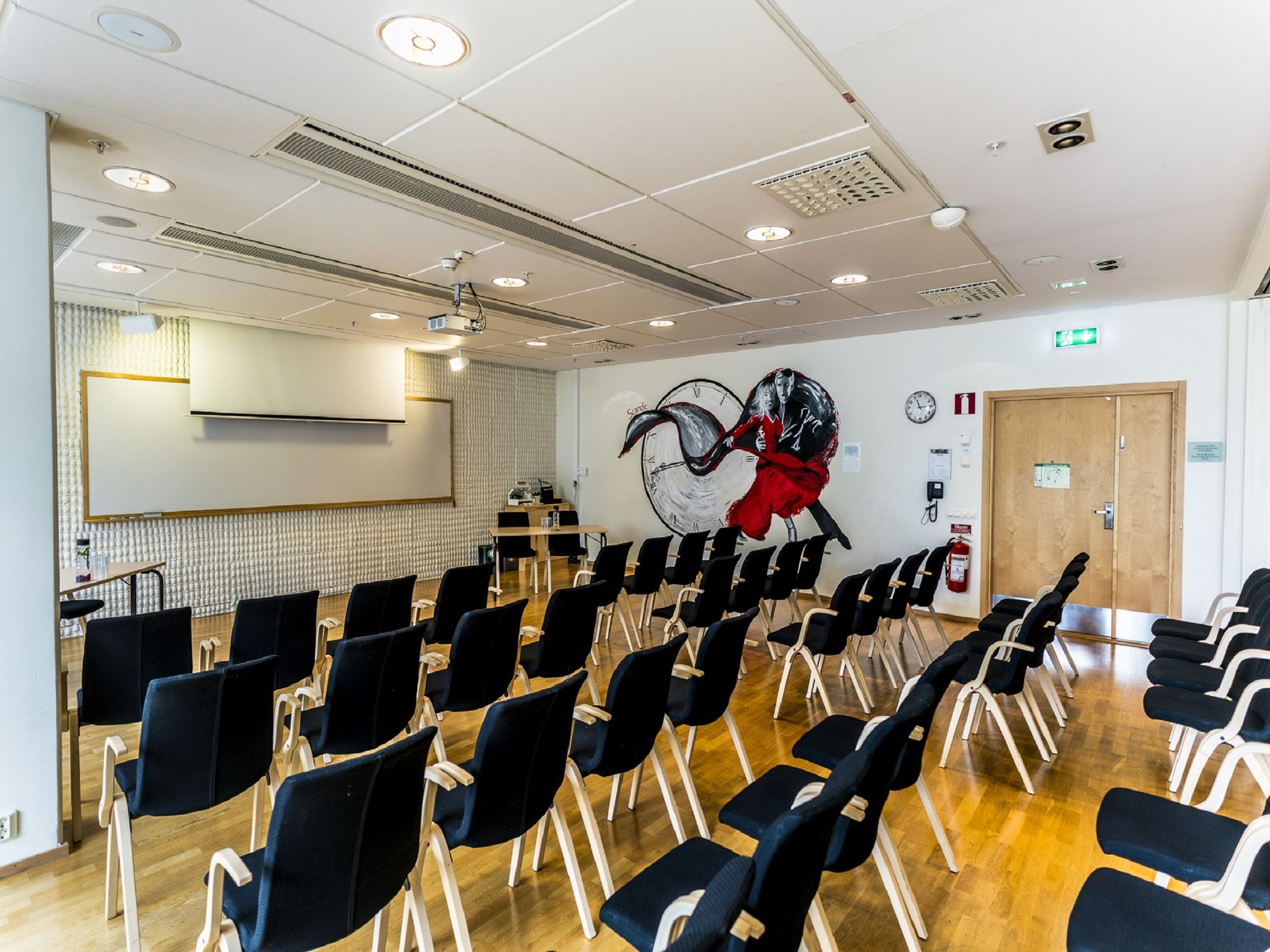 Une salle de conférence avec des rangées de chaises, un tableau blanc, une horloge et un extincteur.