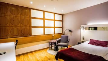 Habitación de hotel con pared de paneles de madera, una cama con manta roja, una silla, una mesita pequeña y una mesita de noche con una lámpara.