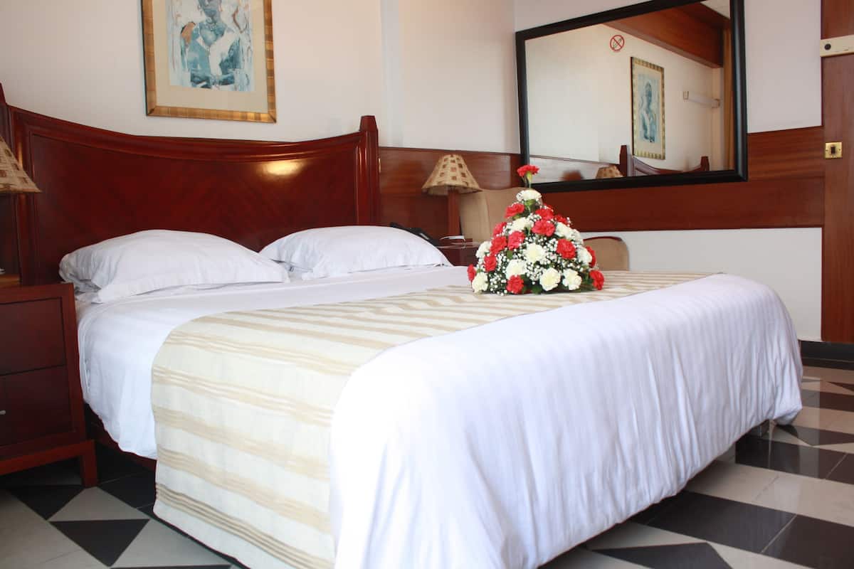 A hotel room with a large bed, a wooden headboard, a nightstand with a lamp, a mirror, and a floral arrangement on the bed.