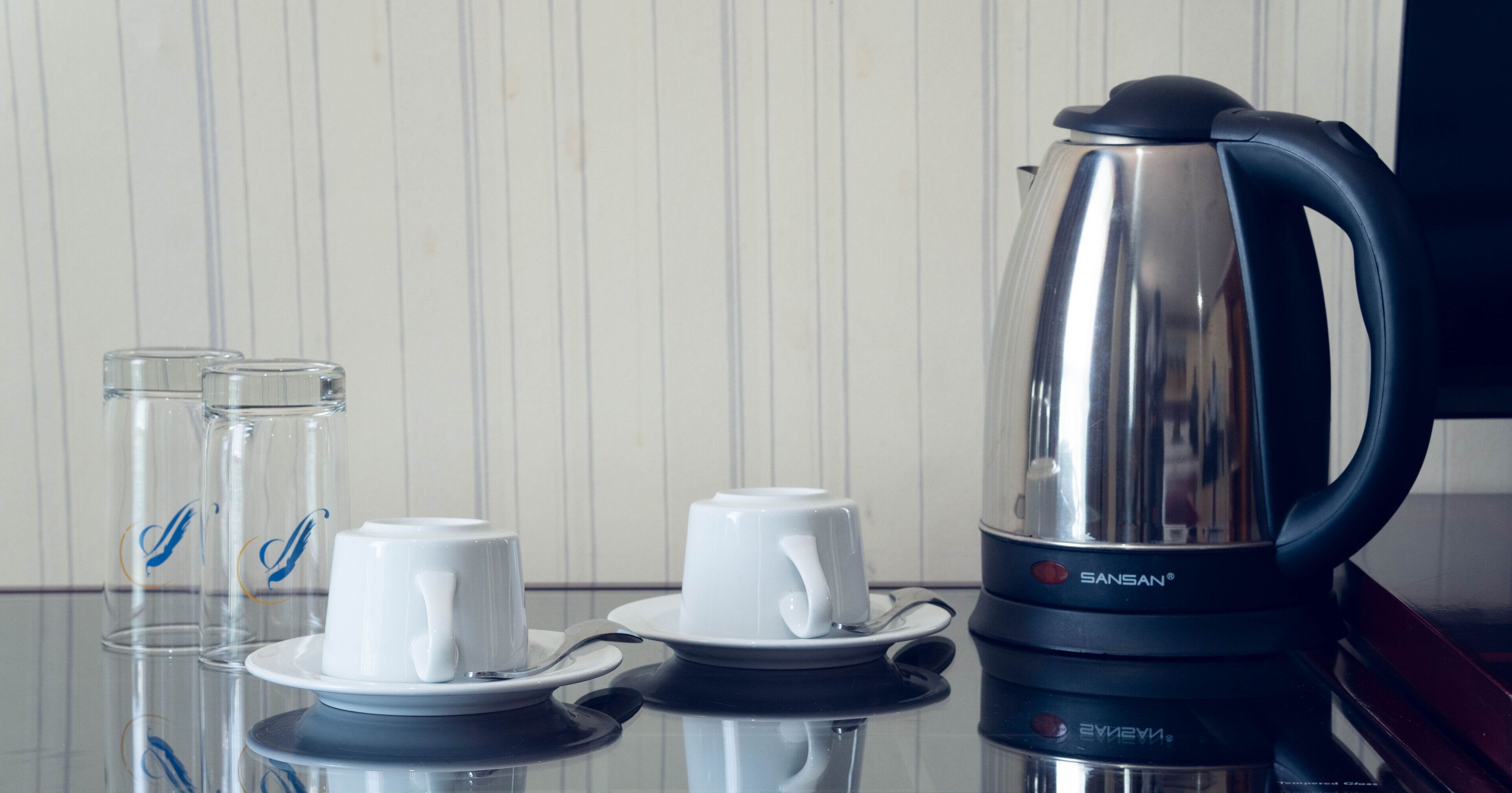 A kitchen area with a stainless steel electric kettle, two white cups with saucers, and two clear glasses with blue designs.