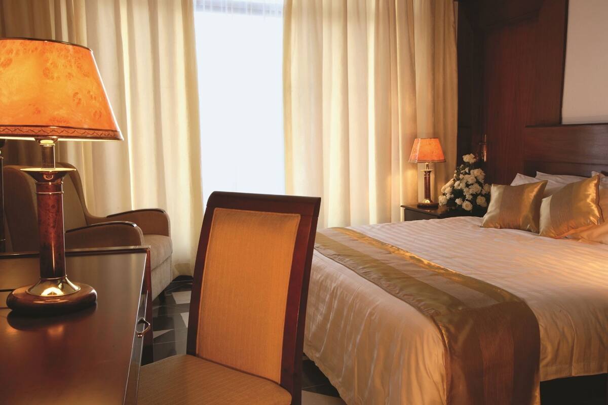 A hotel room with a bed, a desk with a lamp, a chair, a nightstand with a lamp, and curtains.