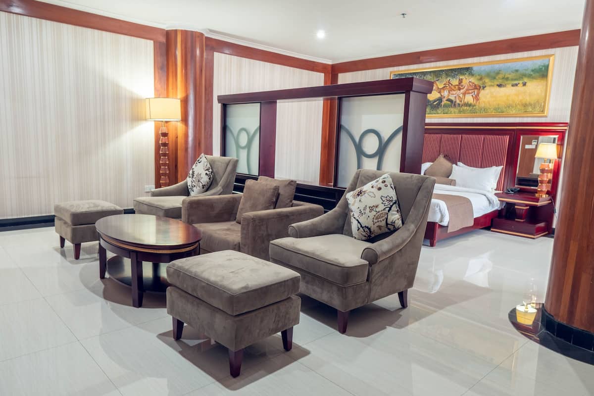 A hotel room with a large bed, a sitting area with sofas and a coffee table, a painting on the wall, and a lamp on the side table.