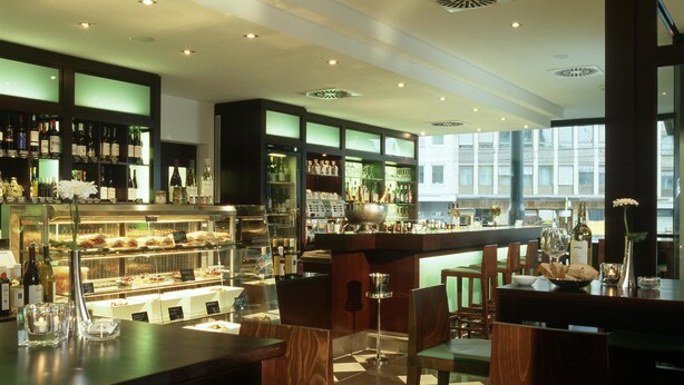 A bar area with wooden furniture, a display case with food, and a view through large windows. - Hotel Lumière an der Messe by ANS (Frankfurt)