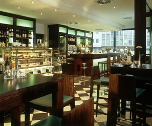 A bar area with wooden furniture, a display case with food, and a view through large windows. - Hotel Lumière an der Messe by ANS (Frankfurt)