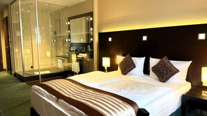 A hotel room with a large bed, a glass-enclosed shower, a bedside table with a lamp, and a headboard with built-in lighting. - Hotel Lumière an der Messe by ANS (Frankfurt)