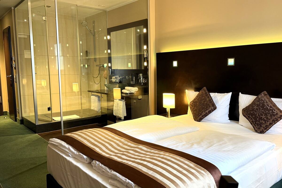 A hotel room with a large bed, a glass-enclosed shower, a bedside table with a lamp, and a headboard with built-in lighting.
