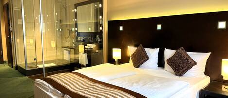 A hotel room with a large bed, a glass-enclosed shower, a bedside table with a lamp, and a headboard with built-in lighting.