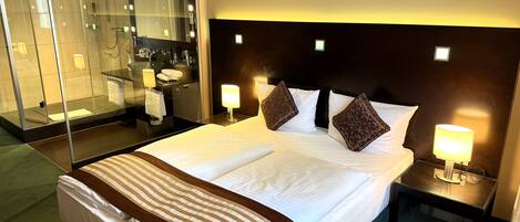 Quarto de hotel com uma cama grande, duas luminárias de cabeceira, mesa de cabeceira, área de chuveiro com box de vidro e um espelho grande.