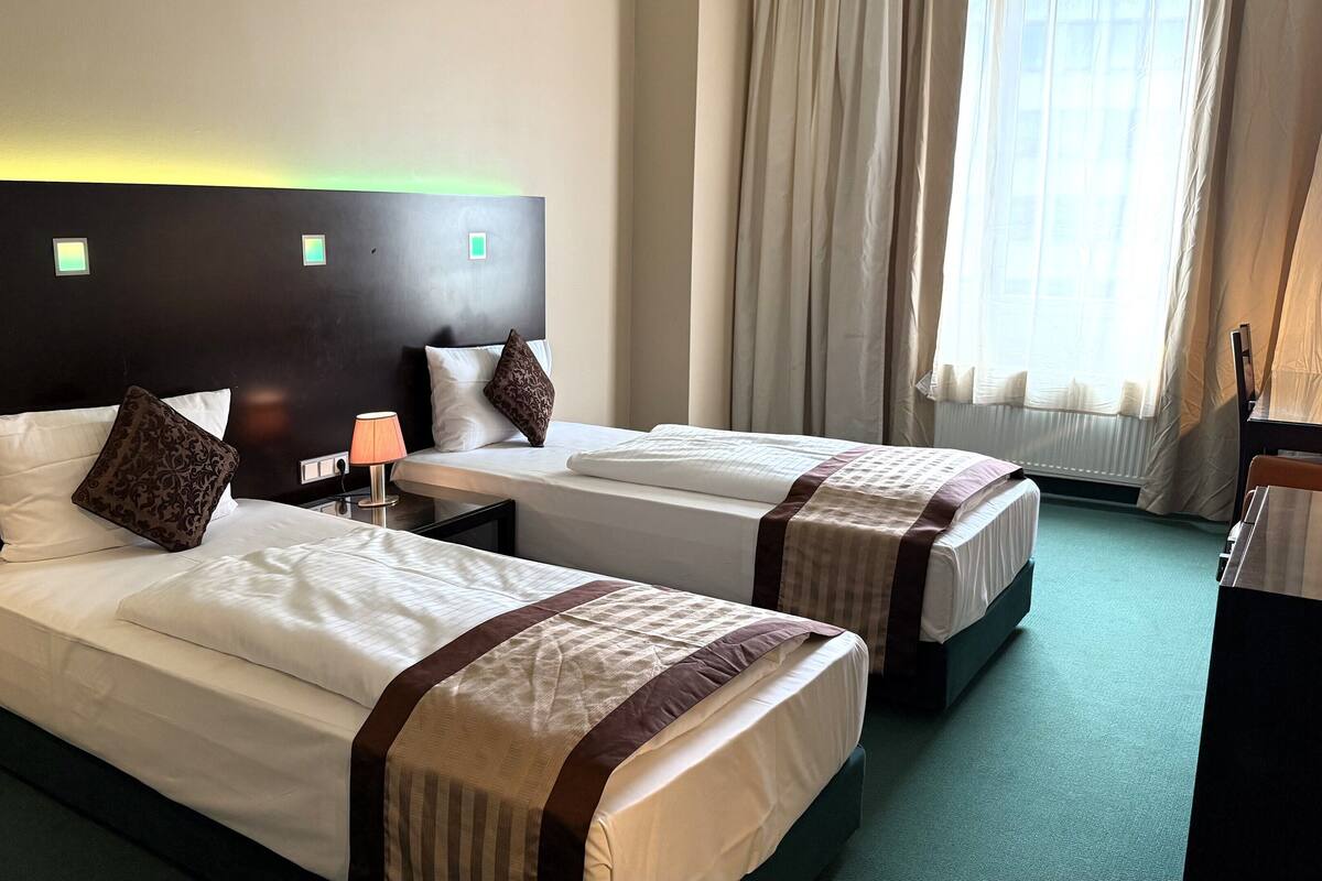 A hotel room with two beds, a nightstand with a lamp, a window with curtains, and a radiator.
