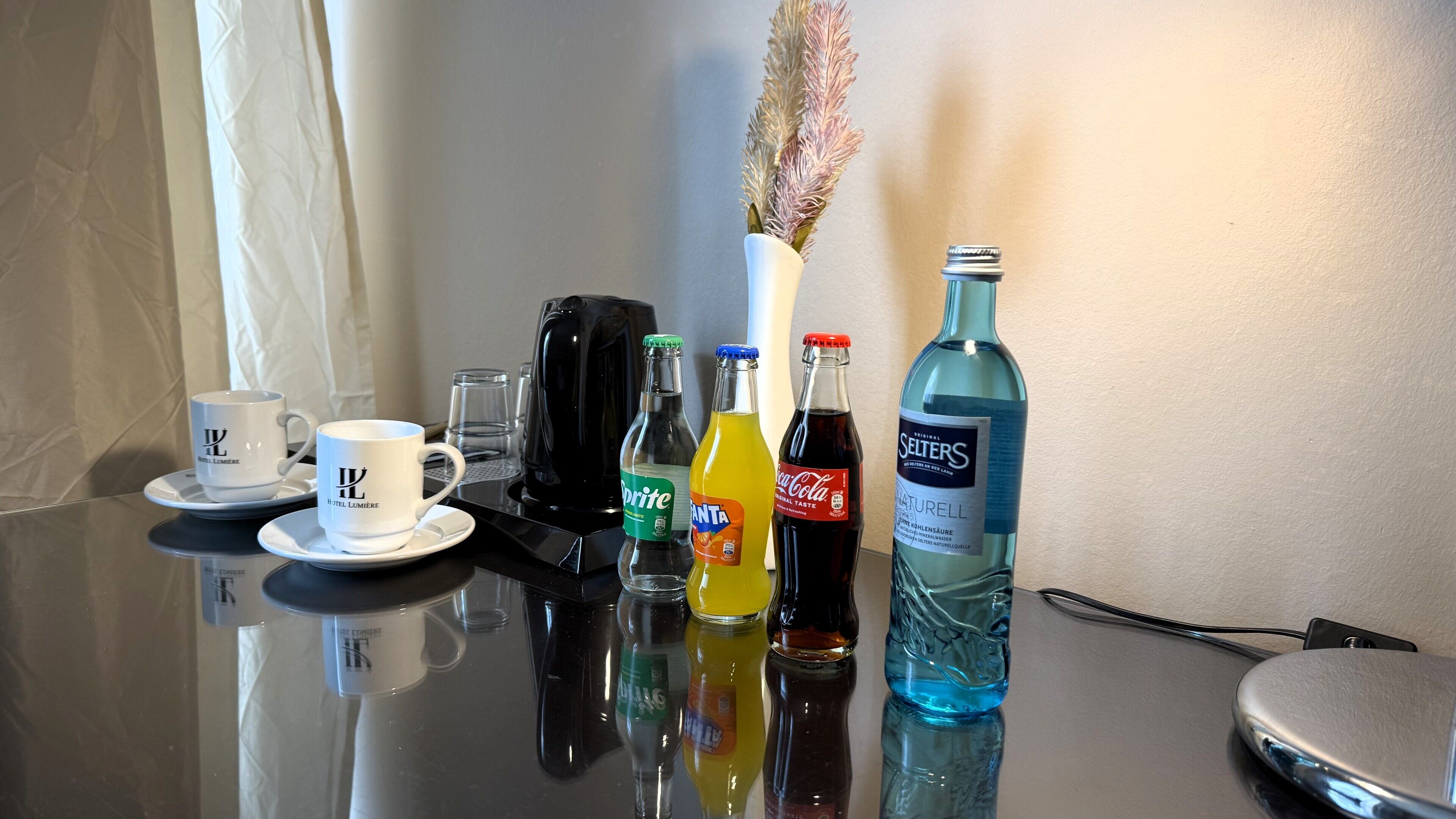 A hotel room table with a coffee cup, a vase with dried flowers, and bottles of beverages.