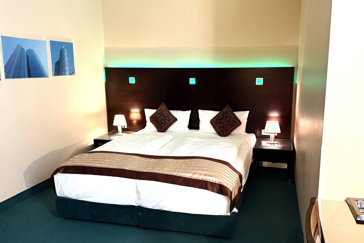 A hotel room with a large bed, two bedside tables with lamps, a headboard with illuminated panels, and a small table with a tray of amenities.
