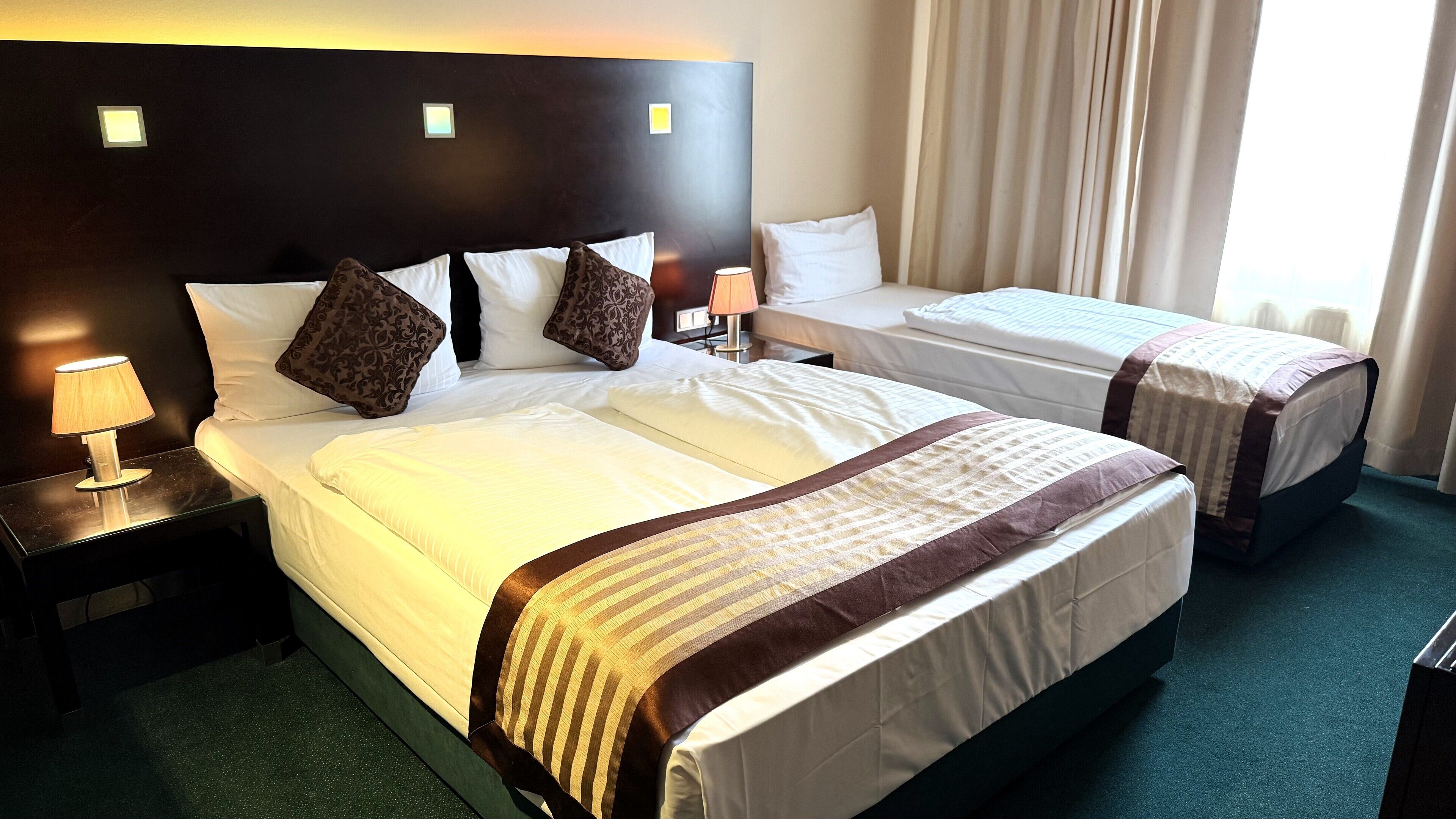 A hotel room with two beds, a nightstand with a lamp, and a window with curtains.