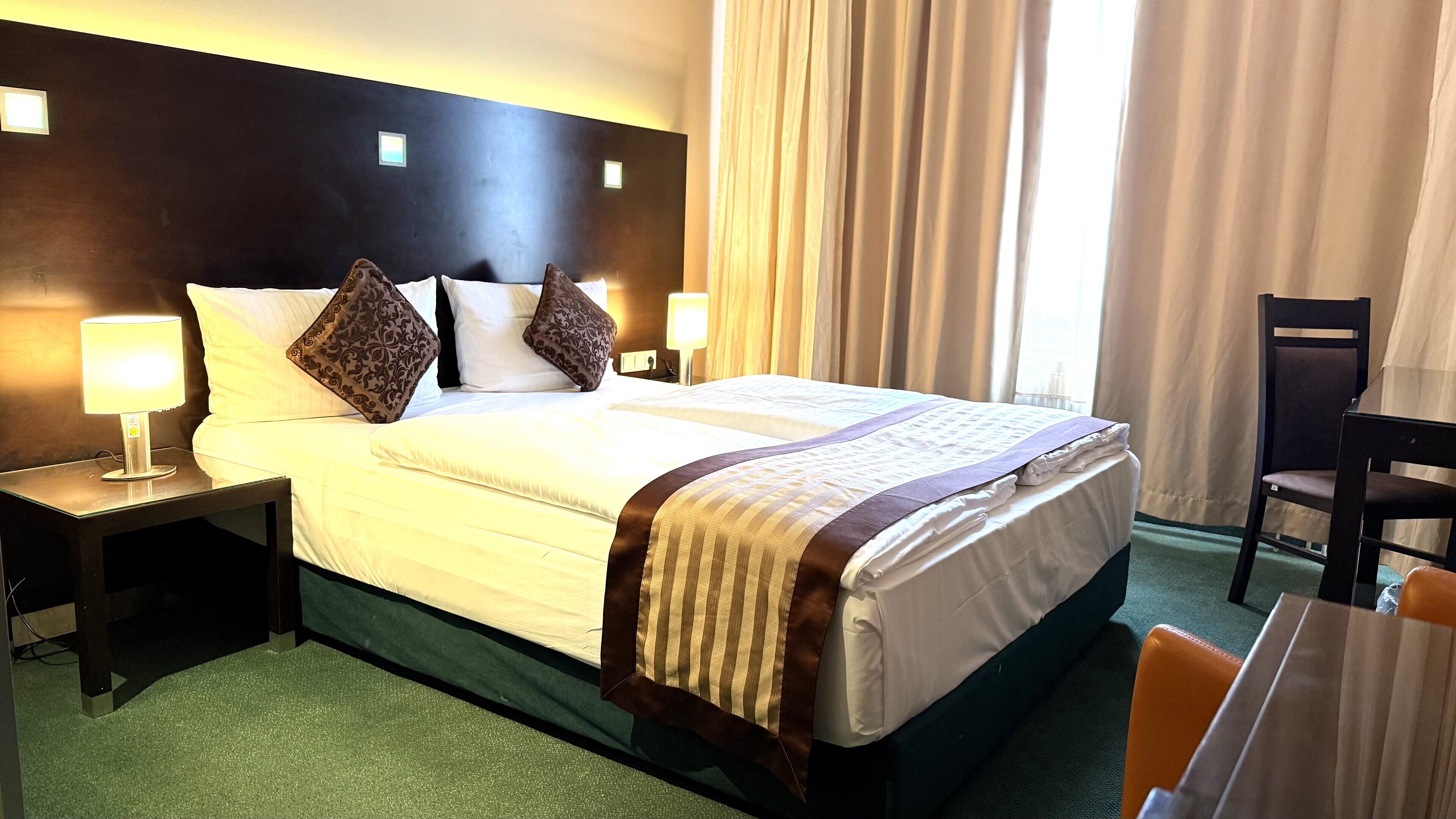 A hotel room with a large bed, two bedside lamps, a chair, and a view of a building through the window.